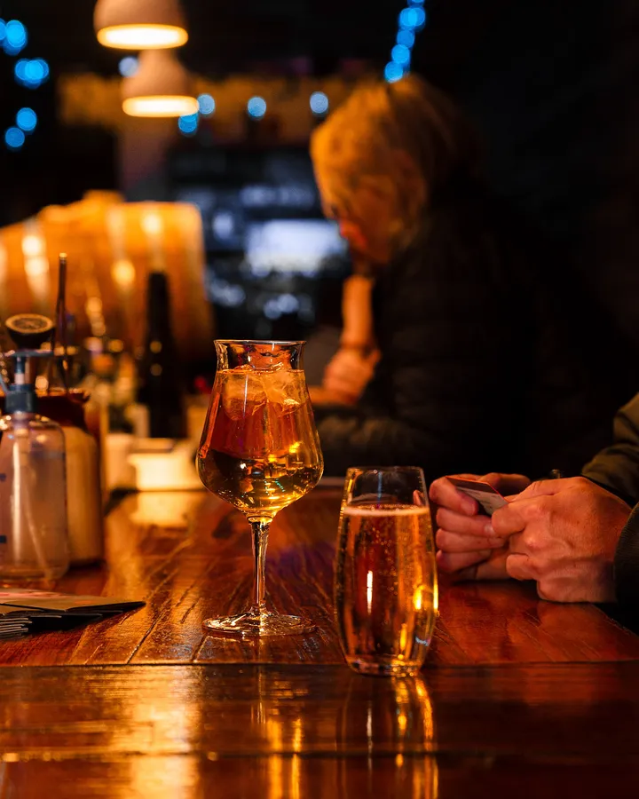 A tulip glass of something golden and a pint on the bar, a guest leaning in behind.