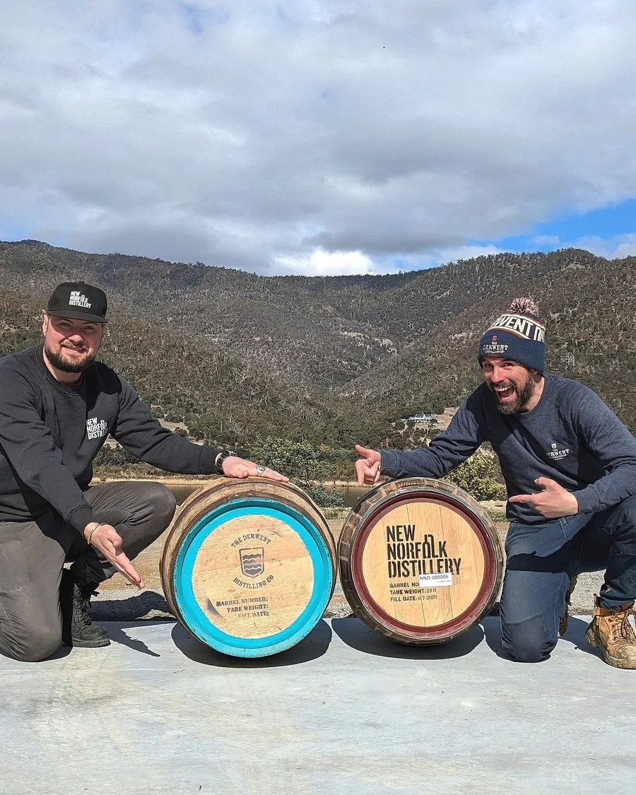 Two people kneeling by a pair of barrels marked New Norfolk Distillery and The Derwent Distilling Co., hills behind them.