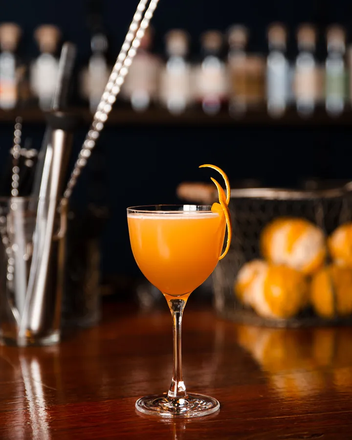 An orange coupe with a twist of peel, a bowl of oranges and bar spoons beside it.