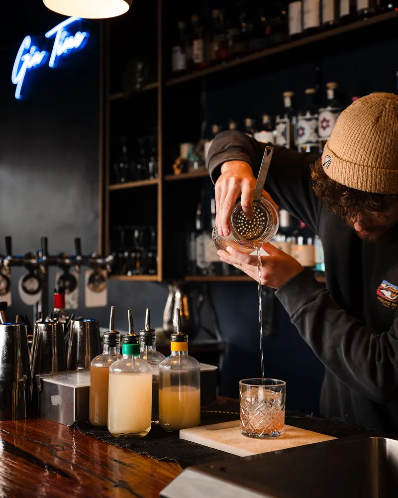 A bartender straining a drink into a tumbler under the blue Gin Time neon.
