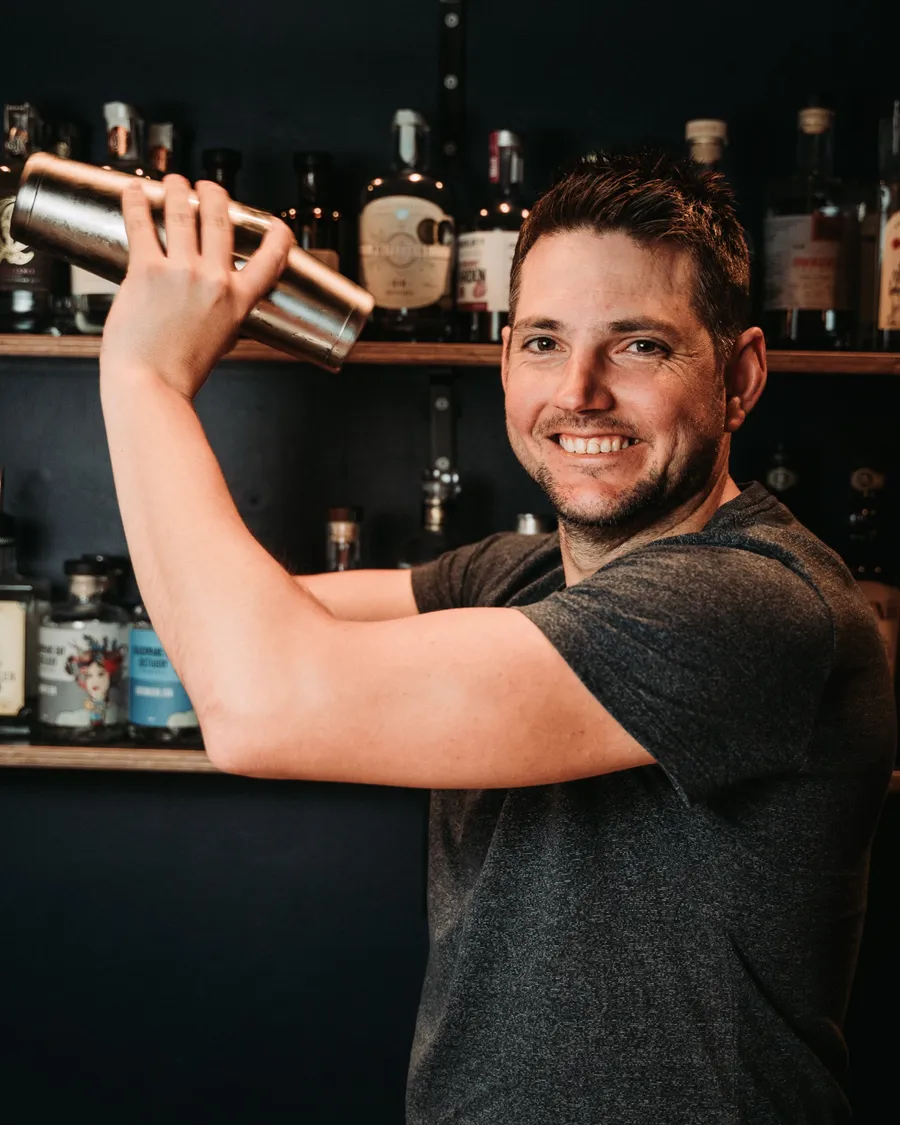 Michael Bernhagen grinning as he shakes a cocktail in front of the back bar's bottle shelves.