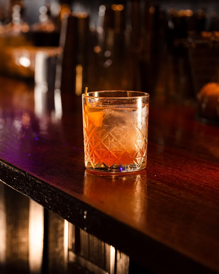 An old fashioned in a cut-glass tumbler, catching the light on the timber bar.