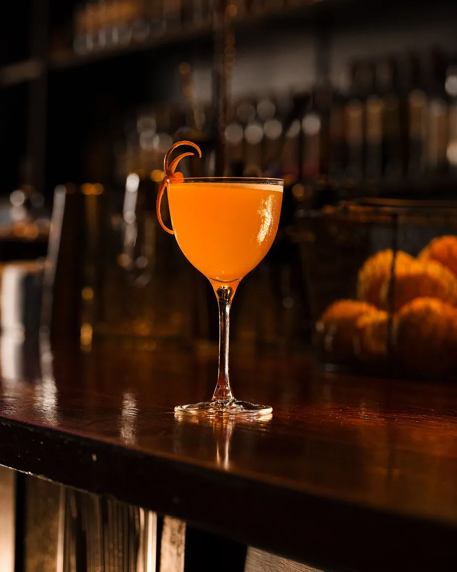 A single orange coupe on the dark timber bar, a curl of peel on the rim and the bottle shelves blurred behind.