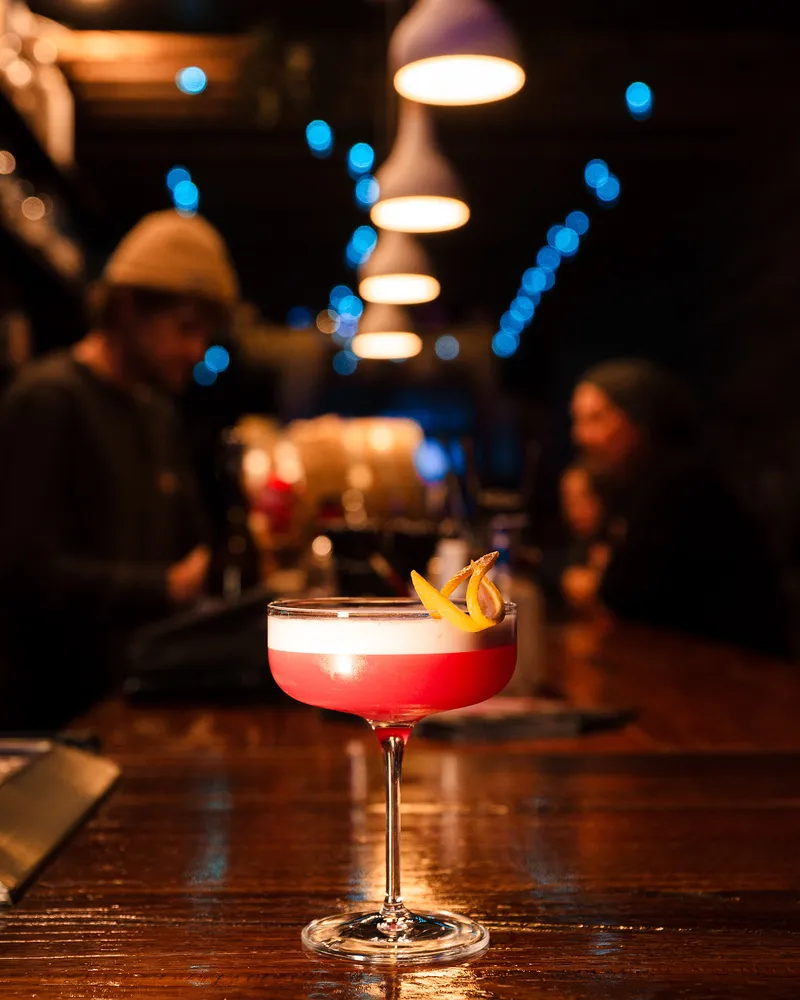 A pink sour with a citrus garnish on the bar, pendant lights and blue string lights glowing down the room.