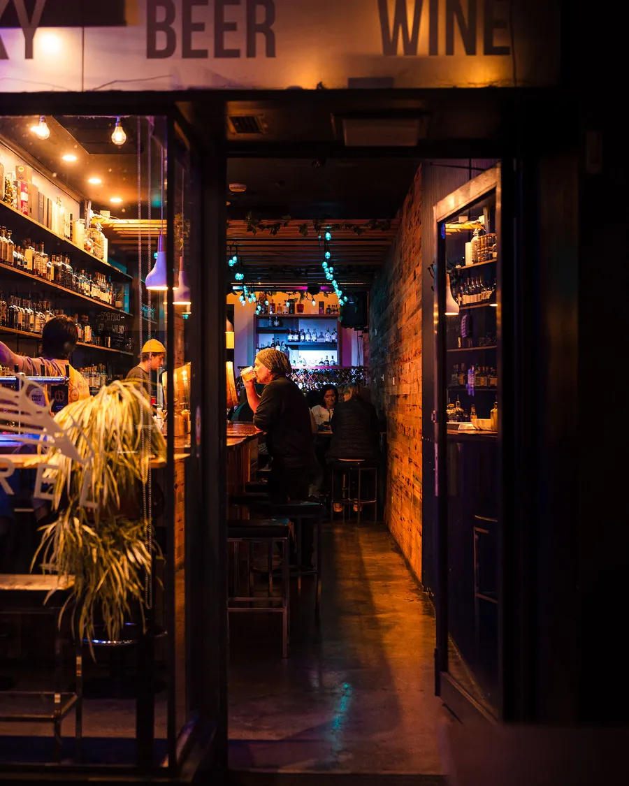 The Barrel Collective's doorway at night, the long bar glowing amber through the glass under signs for whisky, beer and wine.