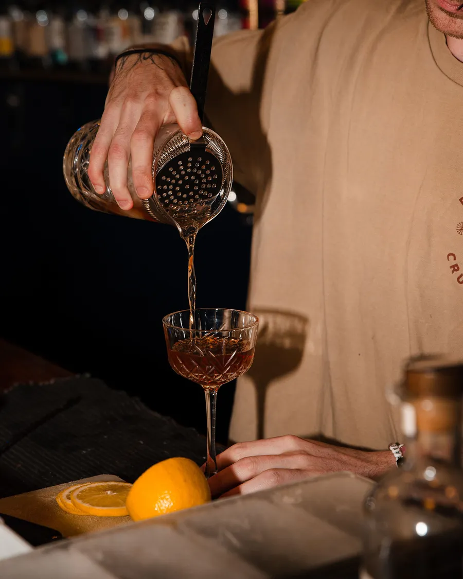 A bartender straining a stirred drink into a cut-glass coupe, a lemon half on the board beside it.