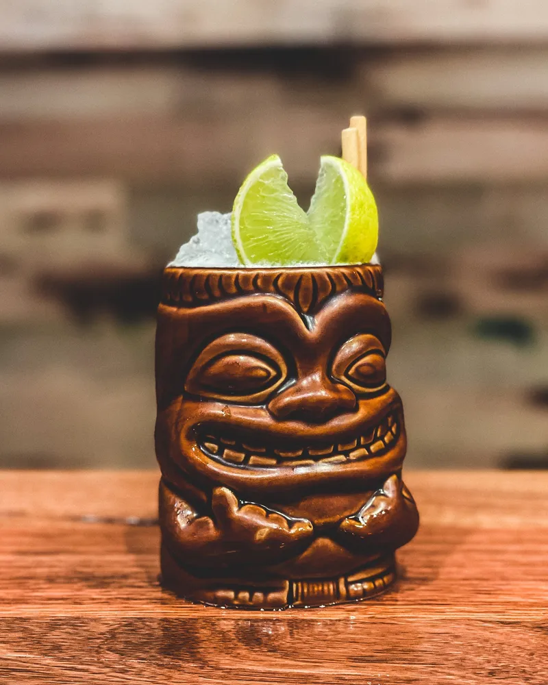 A grinning tiki mug piled with crushed ice and lime wedges on the timber bar.