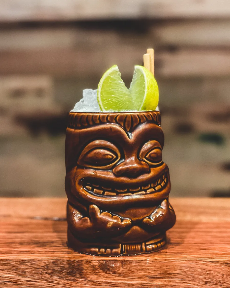 A grinning tiki mug piled with crushed ice and lime wedges on the timber bar.