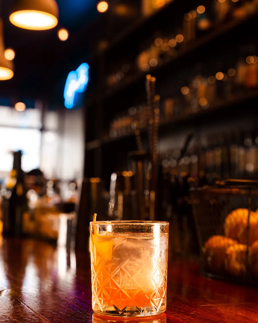 A tumbler with a twist of orange on the bar, blue neon and pendant lights behind.