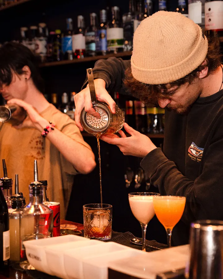 Two bartenders pouring side by side, two coupes waiting on the bar.